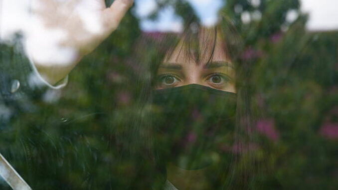 Young woman in black medical mask looking out window, putting hands on window being isolated at home during coronavirus epidemic infection. Thoughtful female with brown eyes observing through window.